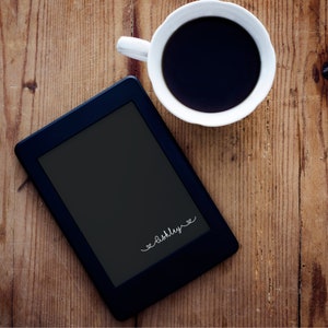 May include: A black e-reader with a white inscription that reads "<3  Ashley <3" on a wooden surface with a white coffee cup filled with black coffee.