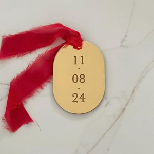 May include: A gold-colored, oval-shaped tag with the numbers "11.08.24" engraved on it. A vibrant red ribbon is attached to the tag. The tag is likely used for gift wrapping or as a decorative element.
