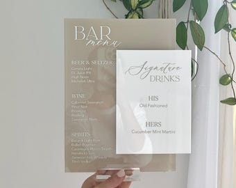 May include: A clear acrylic bar menu with a neutral background. The menu lists beer, wine, and spirits, with signature drinks. The menu is held by a clear acrylic stand. A hanging plant is in the background.
