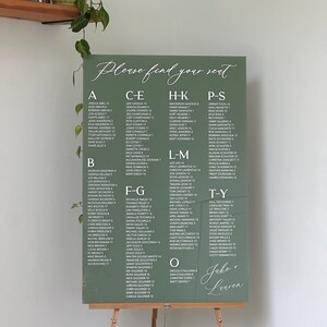 May include: A green seating chart with white text for a wedding reception. The chart is titled "Please find your seat" and lists the names of guests alphabetically by last name. The names are followed by a table number. The chart also includes the names of the bride and groom, "Jake + Lauren".