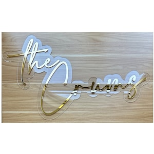 May include: Gold acrylic sign with the words "The Crums" written in a cursive font.