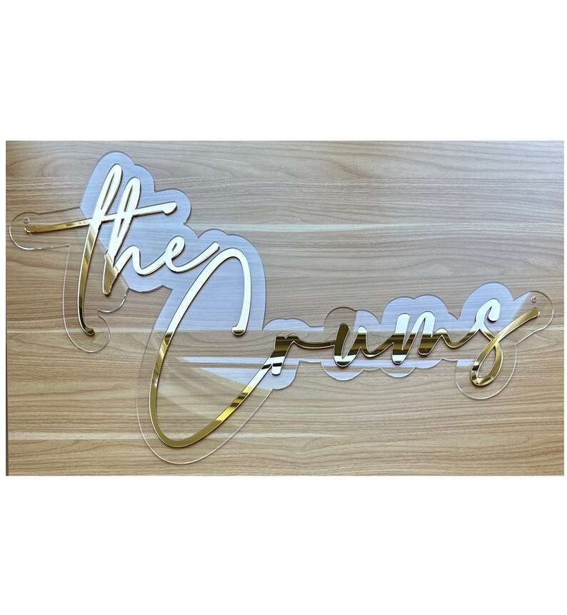 May include: Gold acrylic sign with the words "The Crums" written in a cursive font.