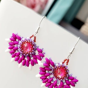 May include: A pair of pink and white beaded earrings with a pink crystal in the center. The earrings are in the shape of a fan and have a silver hook.