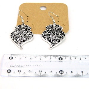 May include: A pair of silver filigree earrings with a heart-shaped design and small clear stones. The earrings are hanging from a ruler, showing they are approximately 2 inches long.
