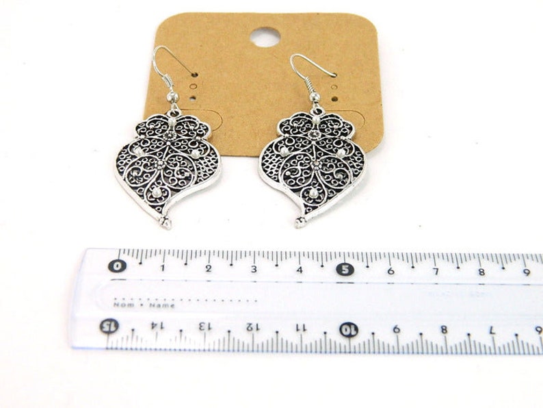 May include: A pair of silver filigree earrings with a heart-shaped design and small clear stones. The earrings are hanging from a ruler, showing they are approximately 2 inches long.
