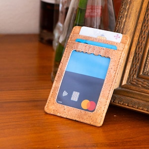 May include: A light brown cork card holder with multiple card slots. The holder contains several credit cards, with a blue card visible through a clear window. The holder is resting on a wooden surface.