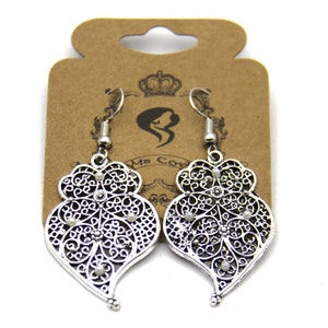 May include: Silver filigree heart-shaped earrings with small white beads. The earrings are hanging from a brown card with the text "Ms. Cow" printed on it.