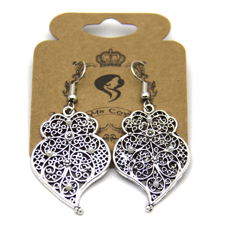 May include: Silver filigree heart-shaped earrings with small white beads. The earrings are hanging from a brown card with the text "Ms. Cow" printed on it.
