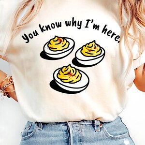 Puede incluir: Camiseta color crema con la frase "You know why I'm here" y un gráfico de tres huevos rellenos. Los huevos tienen relleno amarillo y una pizca de pimentón.