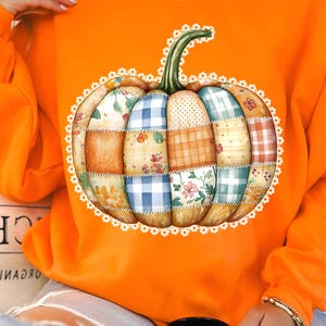 以下が含まれることがあります： パッチワークのかぼちゃのデザインが施されたオレンジ色のスウェットシャツ。かぼちゃは、花柄、ギンガムチェック、無地など、さまざまな生地のパターンで構成されており、白いレースのトリムで縁取られています。スウェットシャツは無地のオレンジ色です。