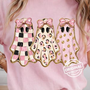 May include: A light pink t-shirt featuring a graphic of three ghosts wearing pink bows. The first ghost has a pink and white checkered pattern, the second has a pink and white leopard print, and the third has pink polka dots. The text ORIGINAL is visible.