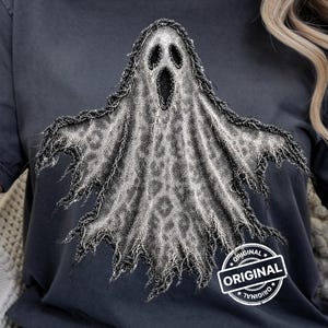 Może przedstawiać: Ciemnoszary t-shirt z krótkim rękawem z dużą grafiką przedstawiającą ducha z teksturowanym wzorem w panterkę. Duch ma postrzępiony, zużyty wygląd. W rogu znajduje się okrągła pieczątka z napisem ORIGINAL.
