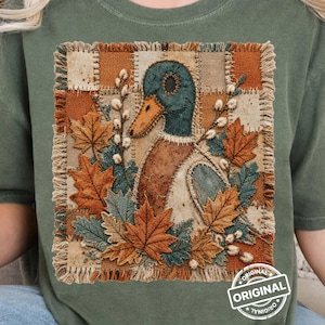Könnte beinhalten: Ein olivgrünes T-Shirt mit einem quadratischen Aufnäher-Design, das eine Stockente, Ahornblätter und Weidenkätzchen zeigt. Der Aufnäher hat ausgefranste Ränder und einen karierten Hintergrund. Ein kreisförmiges Logo mit dem Text ORIGINAL ist zu sehen.