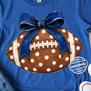 May include: A royal blue short-sleeved t-shirt featuring a graphic of a brown football with white polka dots and a large blue bow on top. The football has white laces and white stripes on the ends. A circular stamp with the text ORIGINAL is visible.