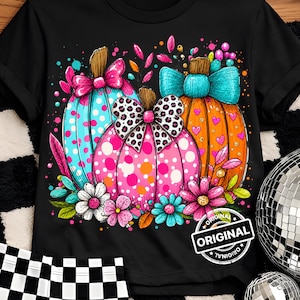 May include: A black short-sleeved t-shirt featuring a graphic of three pumpkins in blue, pink, and orange. The pumpkins are decorated with polka dots, hearts, and bows, accented by colorful flowers. A circular stamp with the text ORIGINAL is visible.