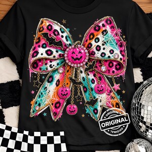 Puede incluir: Una camiseta negra con un gráfico central grande de un lazo con estampado de leopardo rosa. El lazo está decorado con dijes colgantes que incluyen pequeñas calabazas rosas, murciélagos negros, estrellas y telarañas. Una calabaza rosa está en el centro.