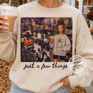 Può includere: Una felpa girocollo beige con una stampa grafica di un carrello della spesa pieno di decorazioni di Halloween, tra cui zucche e teschi, accanto a una persona che indossa una felpa con la scritta Basic Witch on a Budget. Il testo just a few things è stampato sotto la grafica.