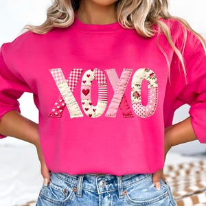 May include: A vibrant pink sweatshirt featuring the letters XOXO. The letters are crafted from diverse patterned fabrics, such as hearts, gingham, and floral designs. The sweatshirt has a crew neck.