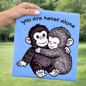 Puede incluir: Un cuadrado azul claro con un gráfico de dos monos abrazándose. Los monos son en blanco y negro con caras sonrientes. El texto "you are never alone" está escrito en escritura negra en la parte superior.