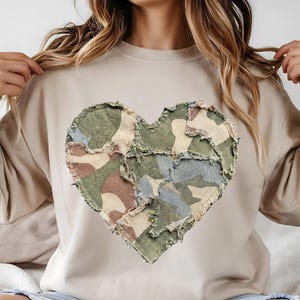 May include: Beige sweatshirt featuring a large heart design made of camouflage fabric patches. The heart is centered on the chest and has a distressed, frayed edge. The sweatshirt has long sleeves and a crew neck.