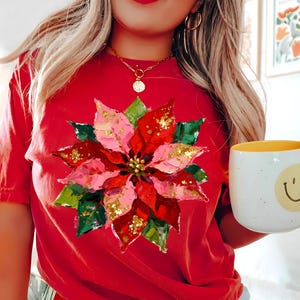 May include: A red t-shirt with a poinsettia flower design in pink, red, green, and gold glitter. The flower is the main feature. A white mug with a yellow interior and a smiley face is held in the hand.