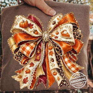 May include: A brown t-shirt featuring a large, multi-layered decorative bow graphic. The bow is composed of ribbons with autumn-themed patterns including pumpkins, acorns, maple leaves, orange gingham, leopard print, and a slice of pumpkin pie.