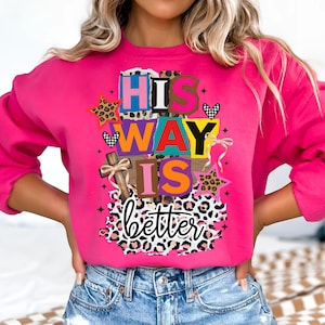 Könnte beinhalten: Ein leuchtend pinkfarbenes Sweatshirt mit dem Schriftzug "HIS WAY IS better" in einem farbenfrohen, mehrschichtigen Design. Das Design umfasst ein Kreuz, Sterne, Herzen und Leopardenmuster-Akzente. Das Sweatshirt ist ein lässiger, bequemer Stil.