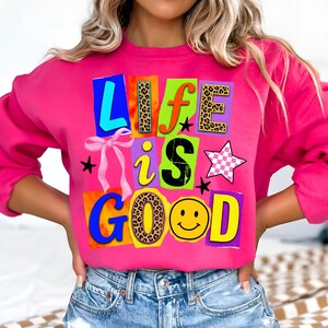 Può includere: Una felpa rosa acceso con la scritta "LIFE IS GOOD" in un design patchwork colorato. Le lettere presentano vari motivi, tra cui stampa leopardata e una faccina sorridente. Un fiocco rosa e stelle completano il design.