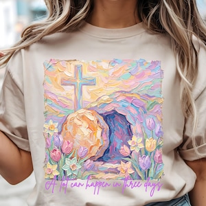 以下が含まれることがあります： ベージュ色のTシャツ。十字架、開いた墓、花が描かれた、カラフルな印象派の絵画が特徴です。「A lot can happen in three days」というテキストが、画像の下部に筆記体で書かれています。