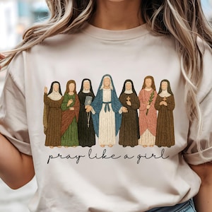 May include: Beige t-shirt featuring an embroidered design of seven figures in religious attire. The figures are in various colors, including brown, green, and blue. The text "pray like a girl" is written in a cursive font below the figures.