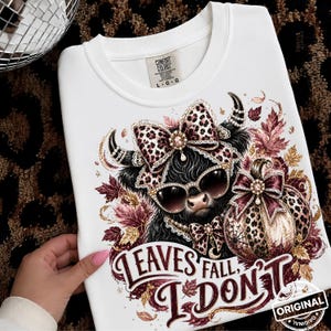 May include: A white crewneck t-shirt featuring a graphic of a black highland cow wearing leopard print sunglasses and a matching bow. The design includes a leopard print pumpkin, autumn leaves, and the text LEAVES FALL, I DON'T.
