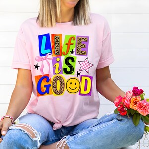 Może przedstawiać: R&oacute;żowa koszulka z napisem "LIFE IS GOOD" w kolorowych, blokowych literach. Litery mają r&oacute;żne wzory, w tym wz&oacute;r w cętki i emotikon uśmiechu. Koszulka jest noszona z niebieskimi dżinsami.