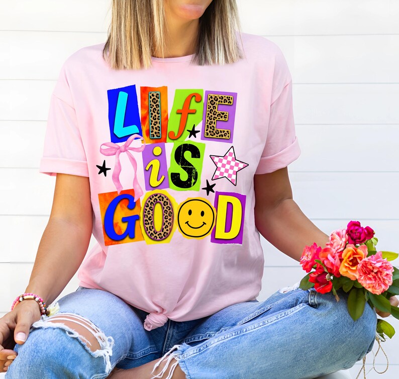 Może przedstawiać: R&oacute;żowa koszulka z napisem "LIFE IS GOOD" w kolorowych, blokowych literach. Litery mają r&oacute;żne wzory, w tym wz&oacute;r w cętki i emotikon uśmiechu. Koszulka jest noszona z niebieskimi dżinsami.