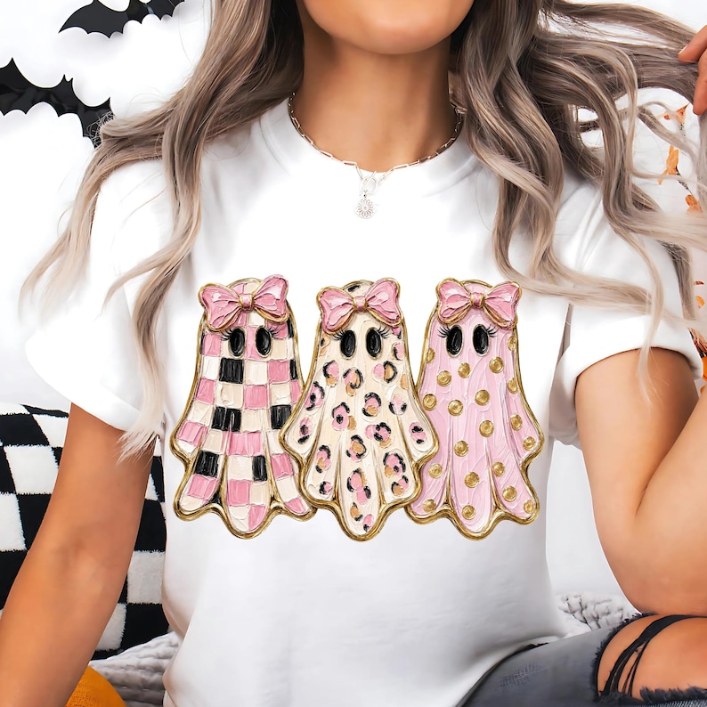 May include: A white short-sleeved t-shirt featuring a graphic of three ghosts wearing pink bows. The first ghost has a pink and white checkered pattern, the second has a leopard print, and the third has pink polka dots with gold accents.
