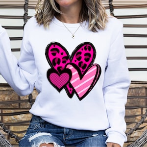 May include: White sweatshirt featuring a pink, black and white heart design. The hearts are layered on top of each other and have different patterns, including leopard print and stripes.