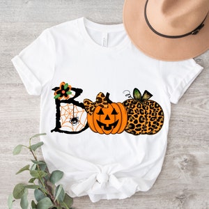 Puede incluir: Camiseta blanca con un gr&aacute;fico negro de "BOO" que presenta una telara&ntilde;a, una calabaza con un lazo y dos calabazas con estampado de leopardo.