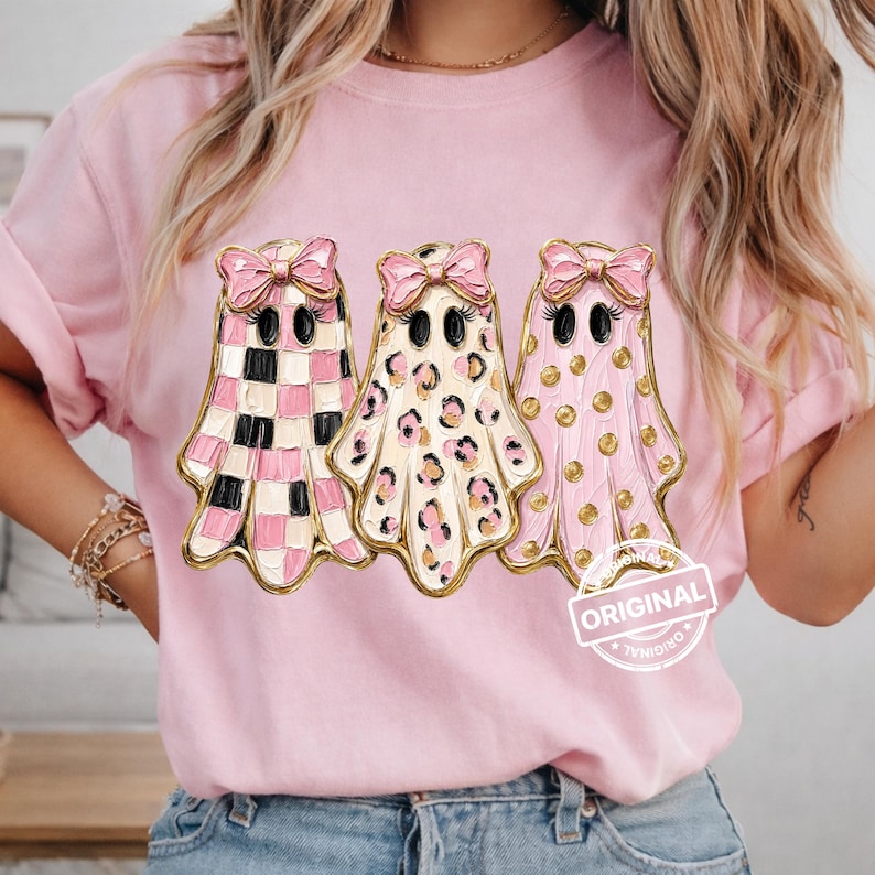 May include: A light pink t-shirt featuring a graphic of three ghosts wearing pink bows. The first ghost has a pink and white checkered pattern, the second has a pink and white leopard print, and the third has pink polka dots. The text ORIGINAL is visible.