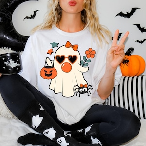 May include: White t-shirt featuring a cute cartoon ghost wearing sunglasses and holding a pumpkin. The ghost has a red nose and a pink bow. There is a spider with a bow on its head below the ghost.