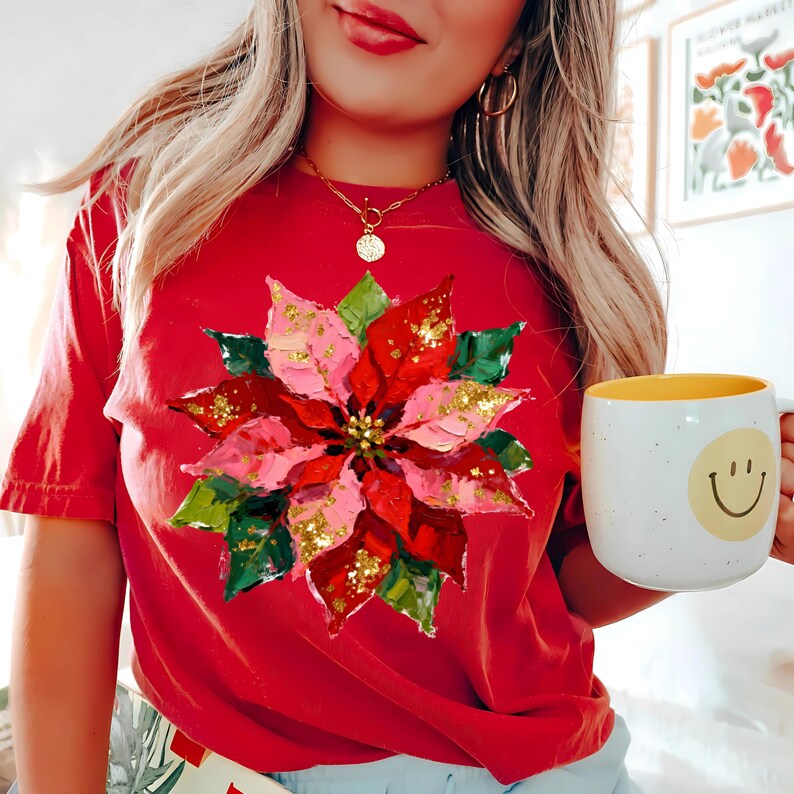 May include: A red t-shirt with a poinsettia flower design in pink, red, green, and gold glitter. The flower is the main feature. A white mug with a yellow interior and a smiley face is held in the hand.