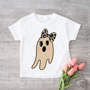 May include: A white t-shirt with a cartoon ghost wearing a leopard print bow. The ghost has a surprised expression with big eyes and an open mouth.