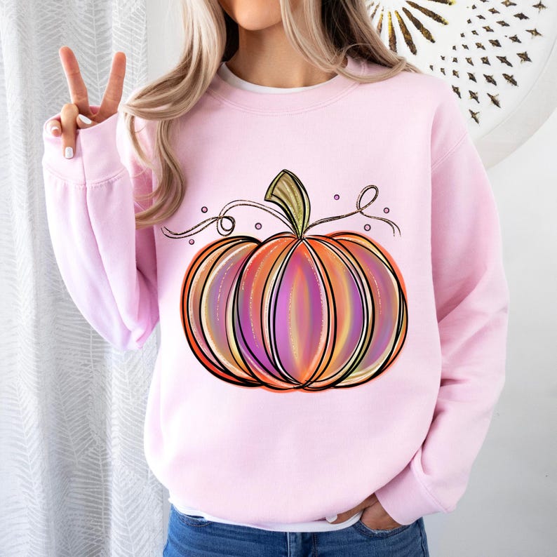 May include: A pink sweatshirt with a colorful, hand-drawn pumpkin design. The pumpkin has orange, yellow, pink, and purple stripes. The pumpkin has a green stem and a vine with leaves.