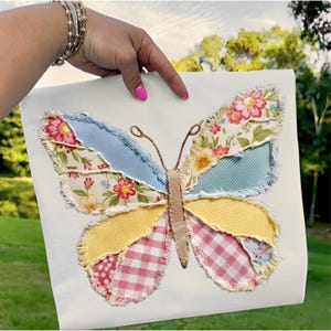 Op de afbeelding: Een kleurrijk vlindervormig kunstwerk van stof. De vleugels zijn samengesteld uit verschillende stoffen met patronen in blauw, geel, roze en bloemenprints. De vlinder heeft een bruin lichaam en antennes, en wordt gepresenteerd op een witte achtergrond.