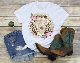Puede incluir: Camiseta blanca con un gráfico de una cabeza de vaca de las tierras altas con acentos florales sobre un fondo de estampado de leopardo. El gráfico está dentro de un marco hexagonal dorado.