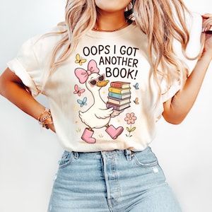 May include: Cream-colored t-shirt with a graphic of a duck wearing sunglasses and a pink bow, carrying a stack of books. The text "OOPS I GOT ANOTHER BOOK!" is above the duck. Butterflies and flowers surround the duck.