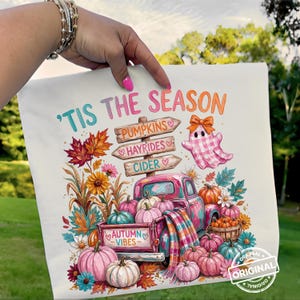 May include: A white t-shirt featuring a colorful autumn graphic with the text 'TIS THE SEASON. The design includes a pink truck, pumpkins, a plaid ghost, and a wooden signpost labeled PUMPKINS, HAYRIDES, and CIDER, with AUTUMN VIBES on the tailgate.