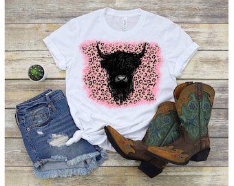 Puede incluir: Camiseta blanca con un gráfico de cabeza de vaca de las tierras altas negras sobre un fondo de estampado de leopardo rosa y negro.