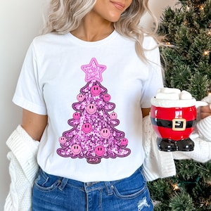 May include: White t-shirt with a pink glitter Christmas tree design. The tree is decorated with pink smiley faces and a pink star on top.