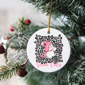 May include: A white ceramic Christmas ornament with a pink and white snowman wearing a pink hat and scarf. The snowman is surrounded by a brown and black leopard print pattern. The text "Snow Cute" is written in pink below the snowman.