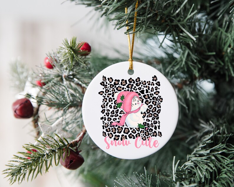May include: A white ceramic Christmas ornament with a pink and white snowman wearing a pink hat and scarf. The snowman is surrounded by a brown and black leopard print pattern. The text "Snow Cute" is written in pink below the snowman.