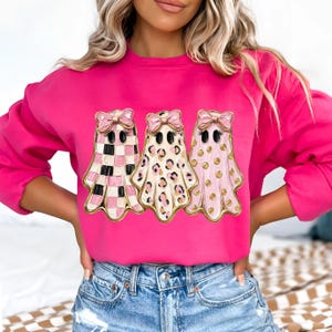 May include: A bright pink long-sleeve sweatshirt featuring a graphic of three ghosts, each wearing a pink bow. The first ghost has a pink and white checkered pattern, the second has a leopard print, and the third has pink polka dots.